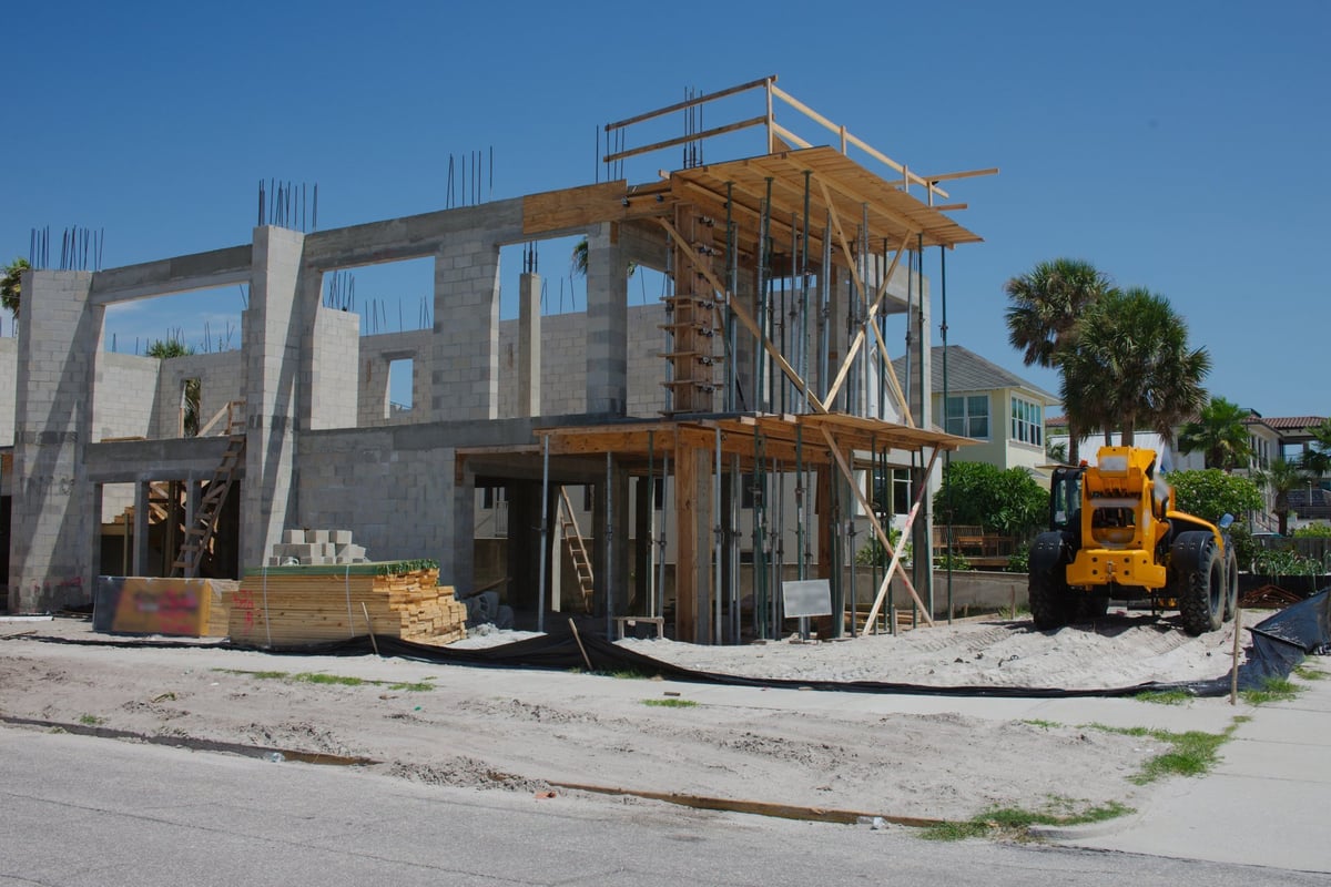 Under a clear sky, a new house structure undergoes construction, showcasing tall walls, open frames, and stacked building materials